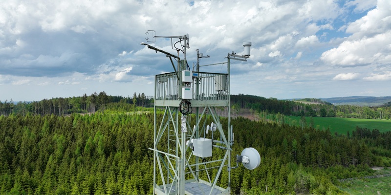 Meteorology tower with pine trees and white clouds in the background