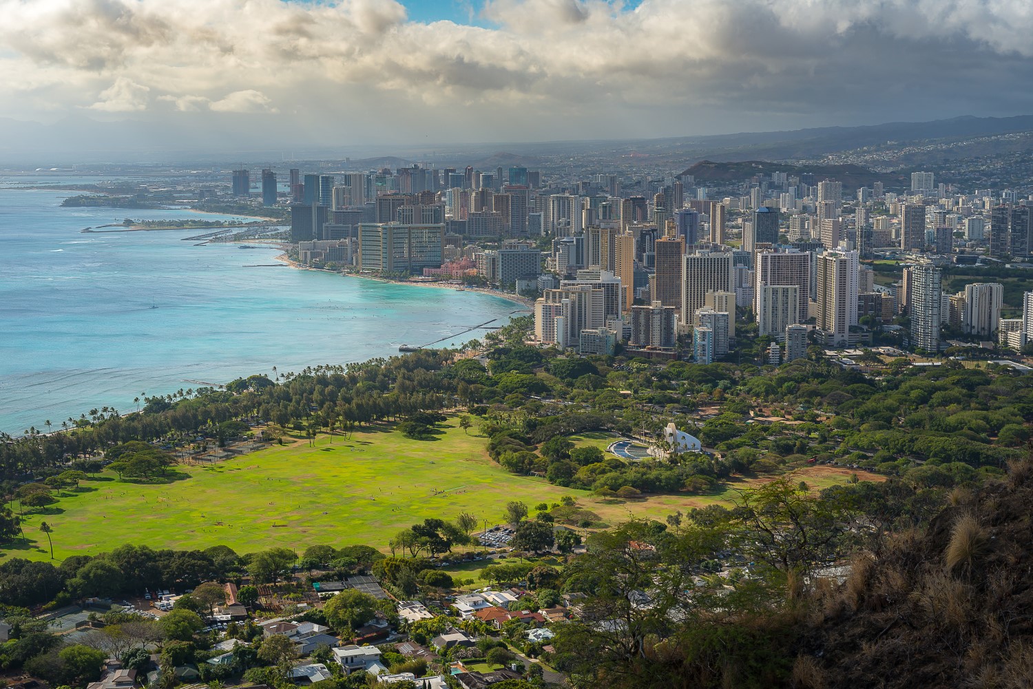 Aerial view of downtown Honolulu skyline, beach and blue waters, with a golf course, amphitheater and residential neighborhood in the foreground and rain clouds in the distance.