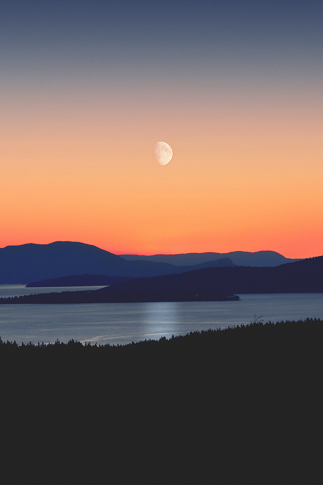 Water with hills underneath a gradient sky and moon