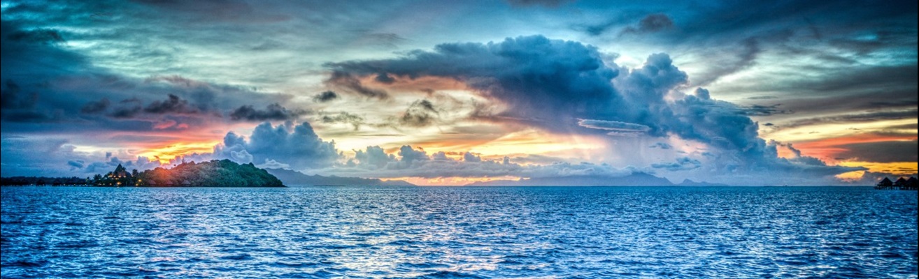 Ocean waves at sunset, with the coast and tall clouds in the distance