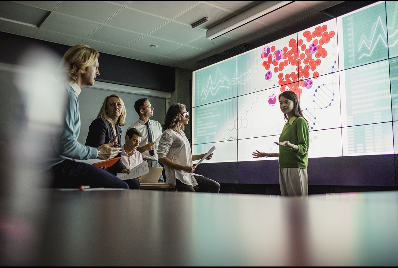 A woman speaks to a group of people in front of a screen A woman speaks to a group of people in front of a screen