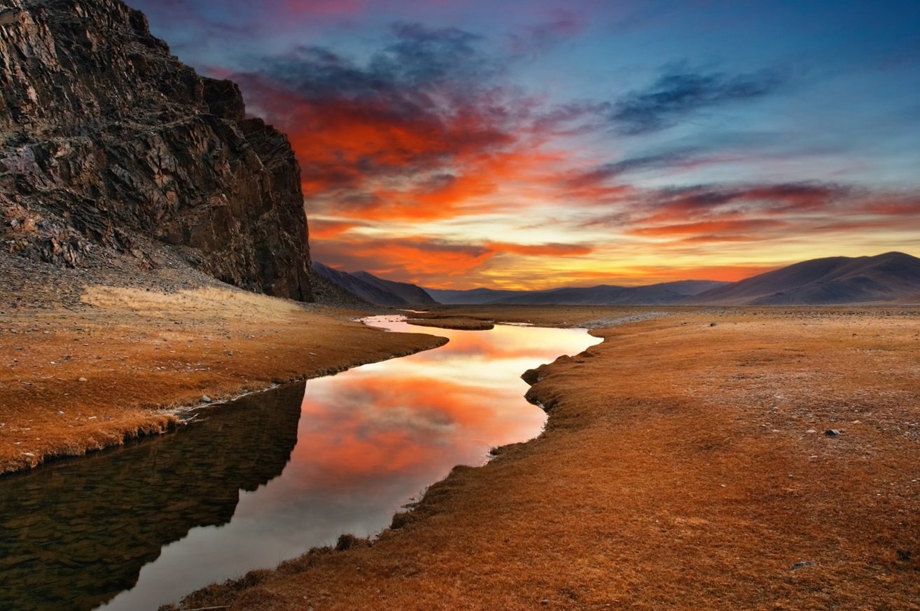 Red cloudy sky and rocky hills reflected in a stream running through a steppe