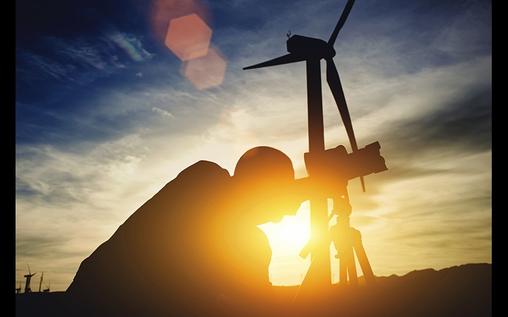 Geodesist using theodolitewhile standing against wind turbine and sunset 