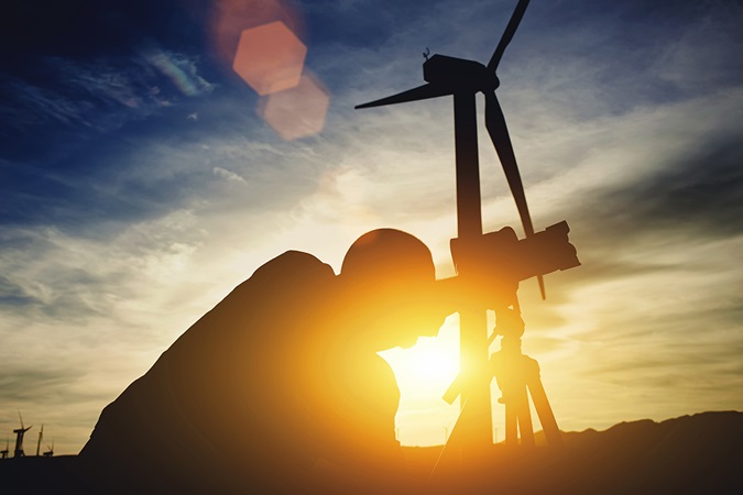 Geodesist using theodolitewhile standing against wind turbine and sunset 
