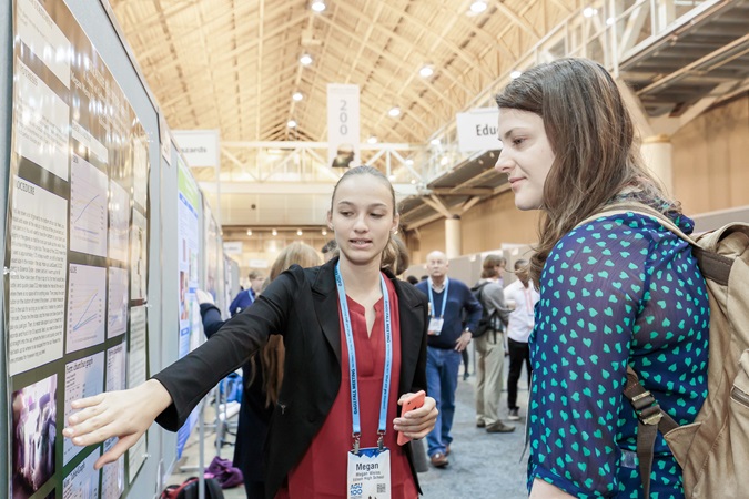 A young woman points to her poster while speaking to another woman