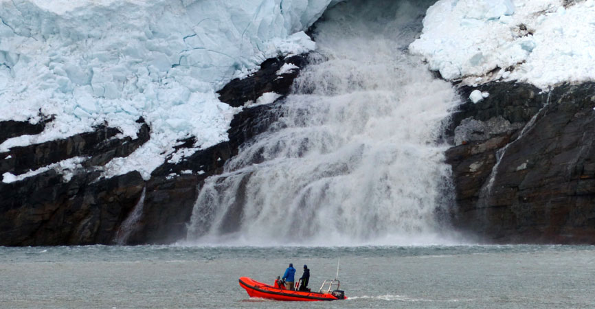 People in red zodiac boat viewing a waterfall emerging from deep snow. Kangerdlugssup Sermerssua fjord, west Greenland