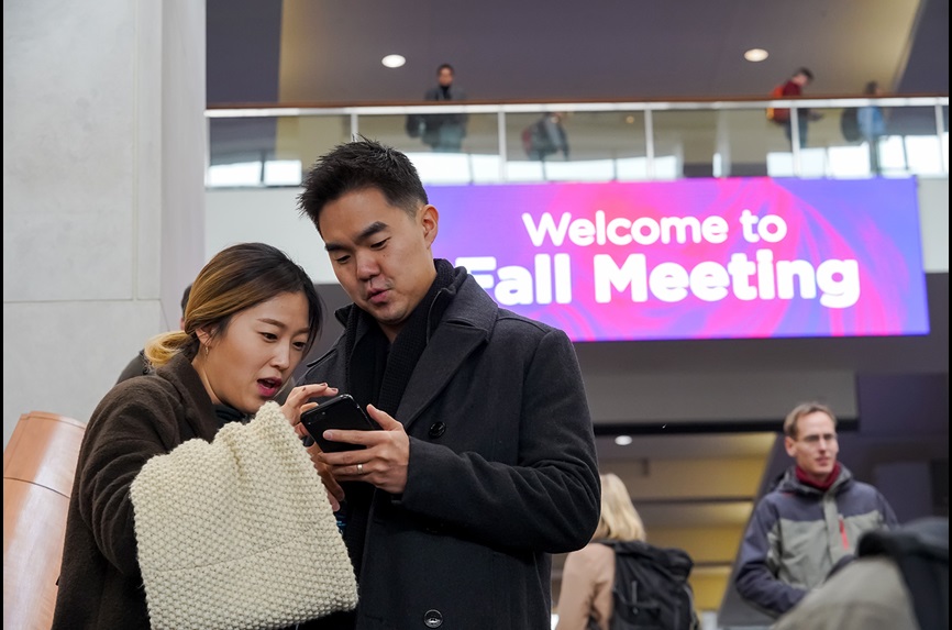 A man and a woman look at a phone with a Welcome to Fall Meeting banner behind them
