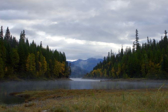 Trees near body of water with foggy mountains in the background. Glacier National Park, Hungry Horse Reservoir, Montana