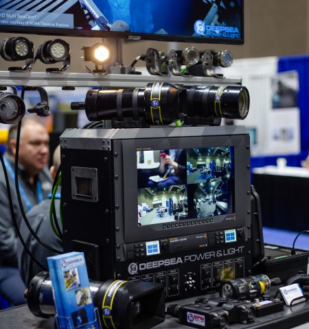 Booth with cameras, lights, a monitor showing 4 video streams from the exhibit hall, and a large screen with underseas image