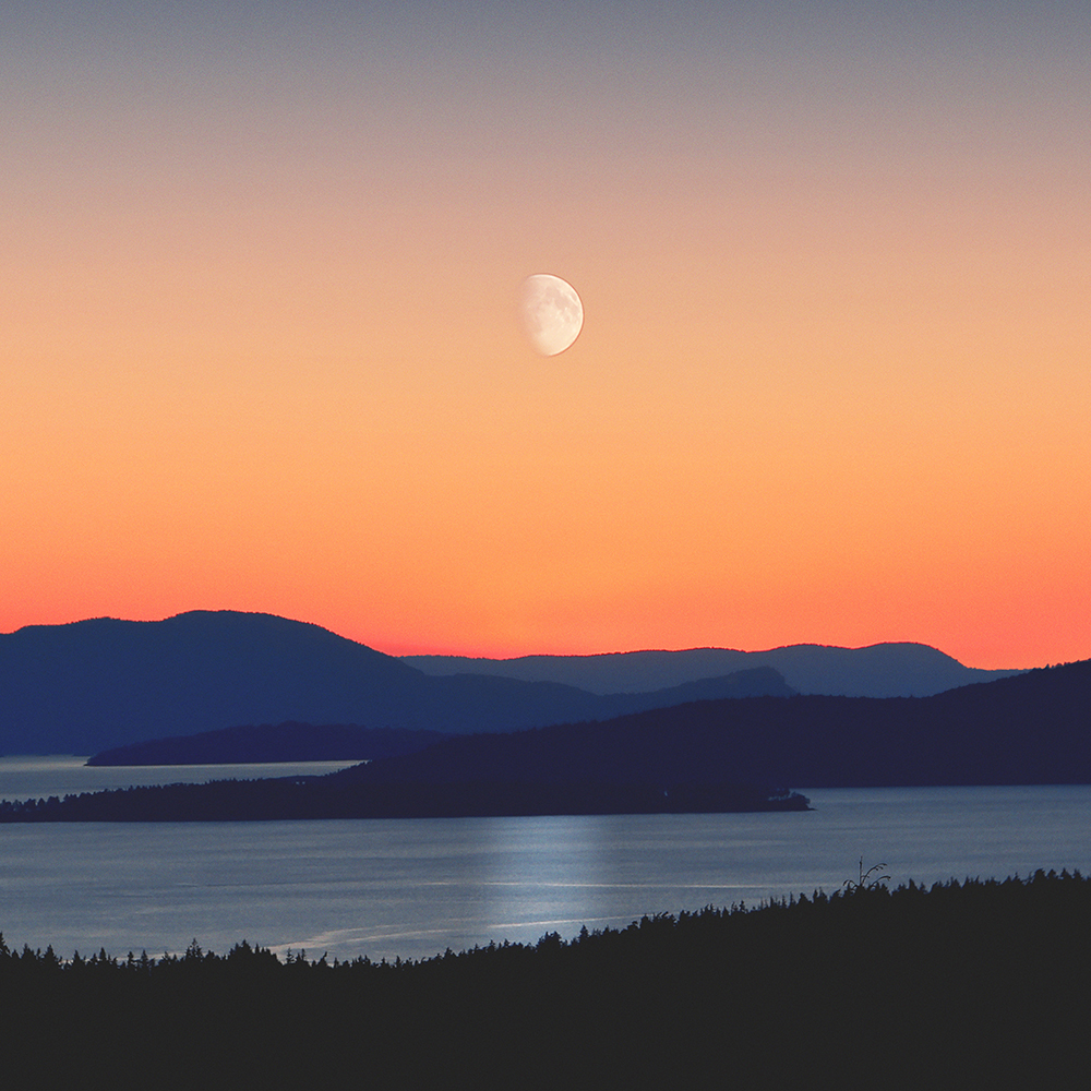 looking out across a lake and mountains with the moon visible at sunset. 