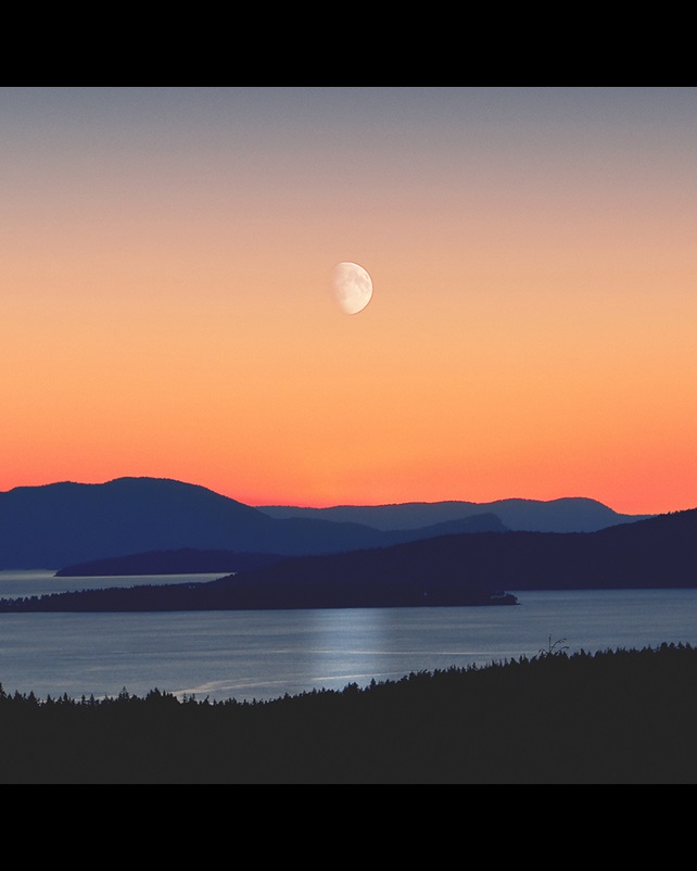 looking out across a lake and mountains with the moon visible at sunset. 