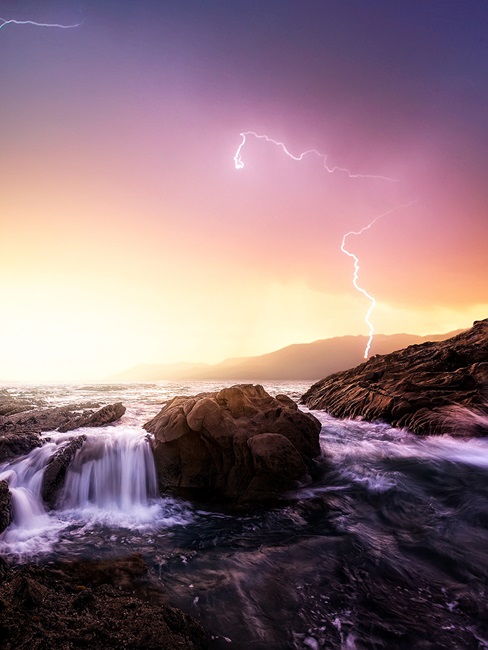 Waves crashing over rocks with lightning strike in background